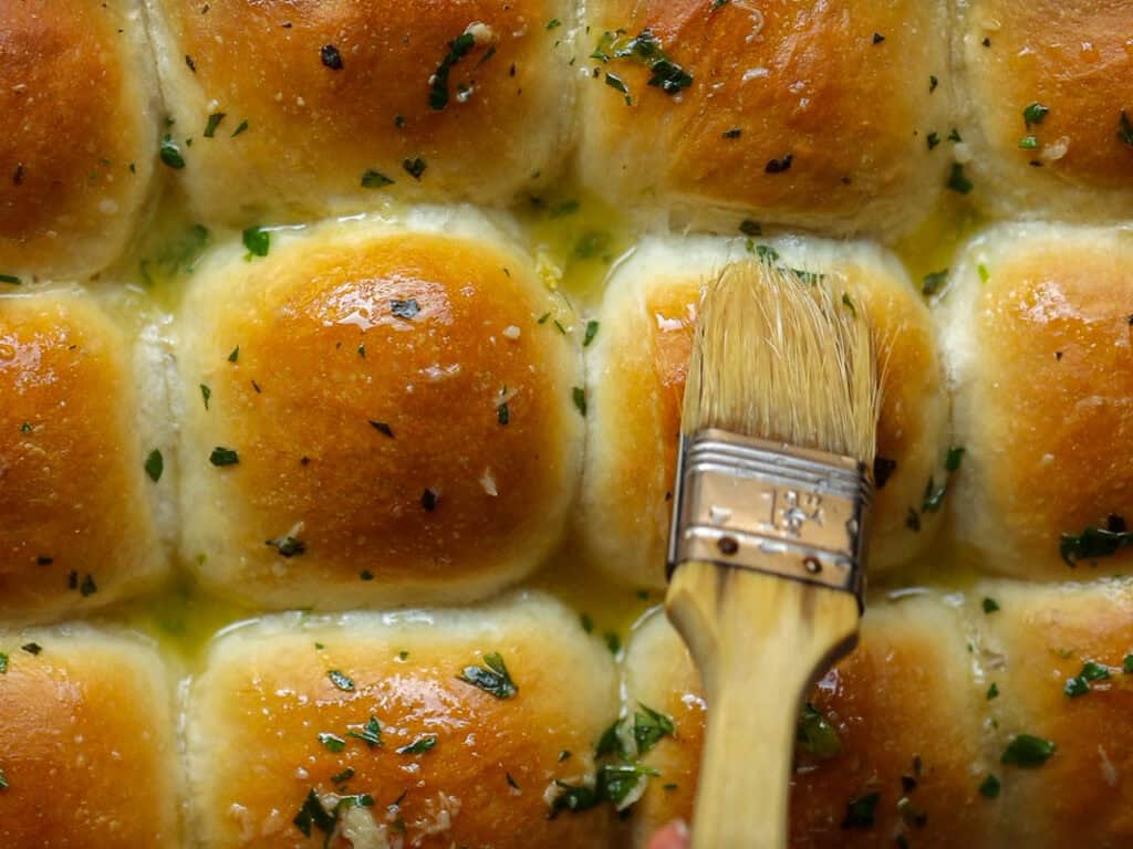 A close-up of golden-brown vegan dinner rolls being brushed with melted garlic butter, garnished with chopped parsley.